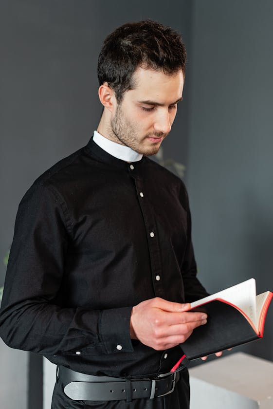 A young male priest dressed in black reads a religious book indoors, embodying spirituality and contemplation.