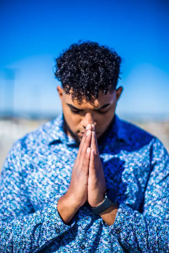 Adult man with curly hair praying outdoors under a clear blue sky, wearing a patterned shirt.