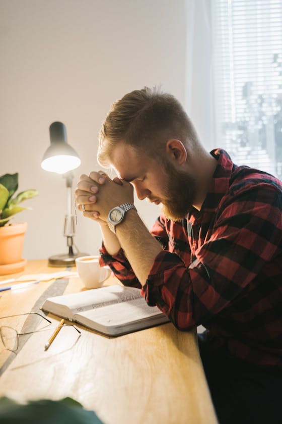 Bearded man in plaid shirt praying with closed eyes at desk with Bible under soft indoor lighting.