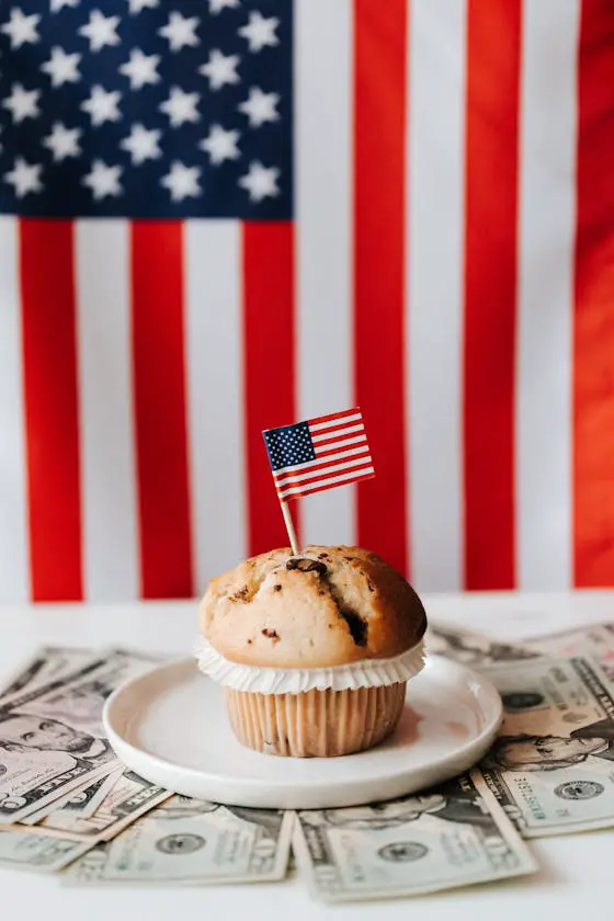 Muffin with US flag toothpick on plate surrounded by dollar bills and an American flag background.