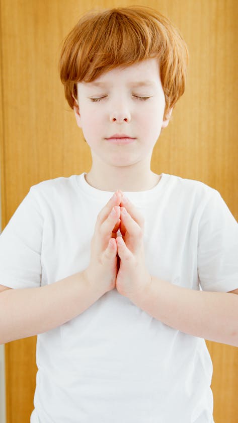 Young red-haired child in meditation pose with eyes closed, wearing white shirt indoors.